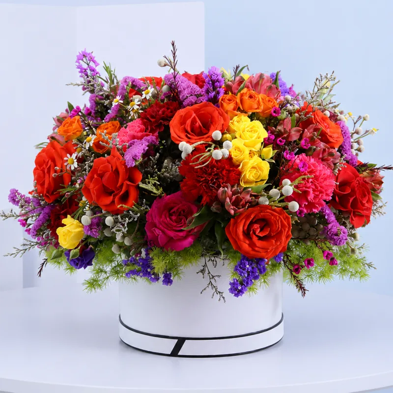 vibrant-bloom-floral-hat-box Vibrant Bloom Floral Hat Box featuring fresh red roses, yellow spray roses, pink carnations, alstroemeria, brunia, purple and blue statice, pink wax flowers, white tanacetum, and asparagus foliage beautifully arranged in a premium white hat box for gifting and special occasions.