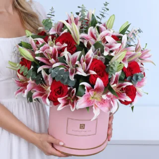 Scarlet Rose Symphony featuring red roses, pink Oriental lilies, and Baby Blue eucalyptus in a pink hatbox.