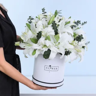 Serene White Lilies Box featuring white Oriental lilies and Baby Blue eucalyptus arranged in a white hatbox.