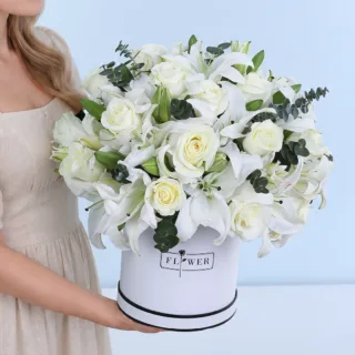 Snowy Floral Whispers featuring white roses, white Oriental lilies, and Baby Blue eucalyptus in a white hatbox.