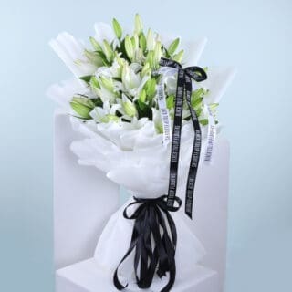 BTF Home 25 A large bouquet of pristine white lilies, elegantly wrapped in white tissue paper and tied with a black ribbon.