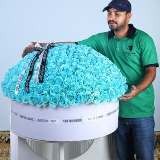 BTF Home 57 A man in a green polo shirt gestures towards a very large, round floral arrangement made entirely of vibrant aqua blue roses in a white box.